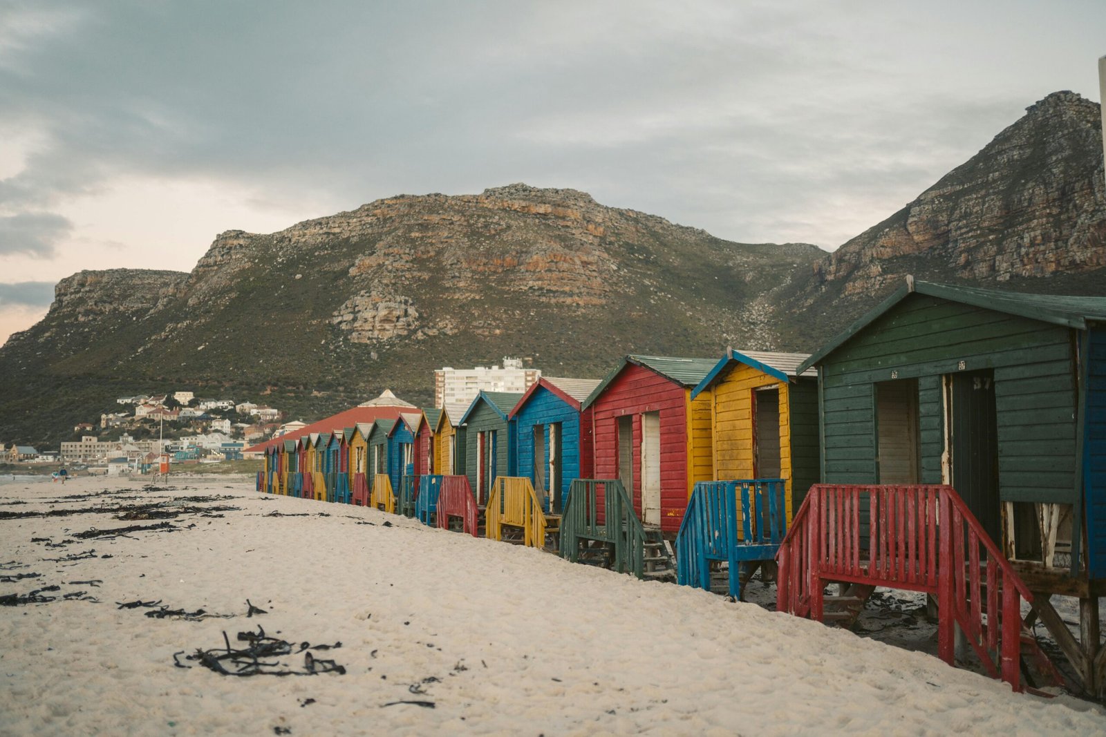 Home Vibrant wooden beach huts on sandy Muizenberg Beach with mountains in the background.
